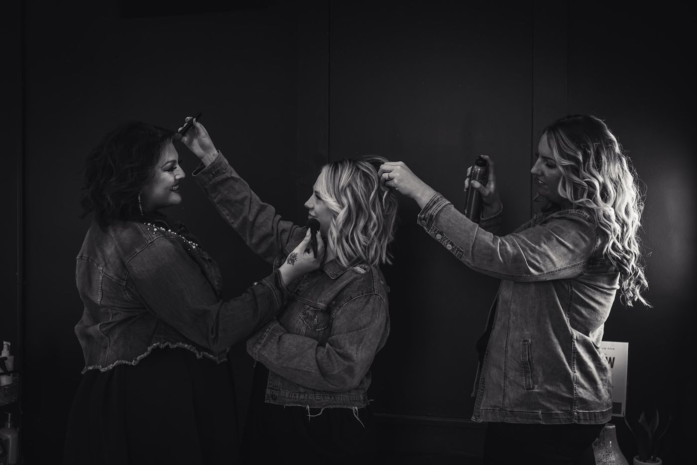 Black and white photo of three stylists in denim jackets styling a client's hair together